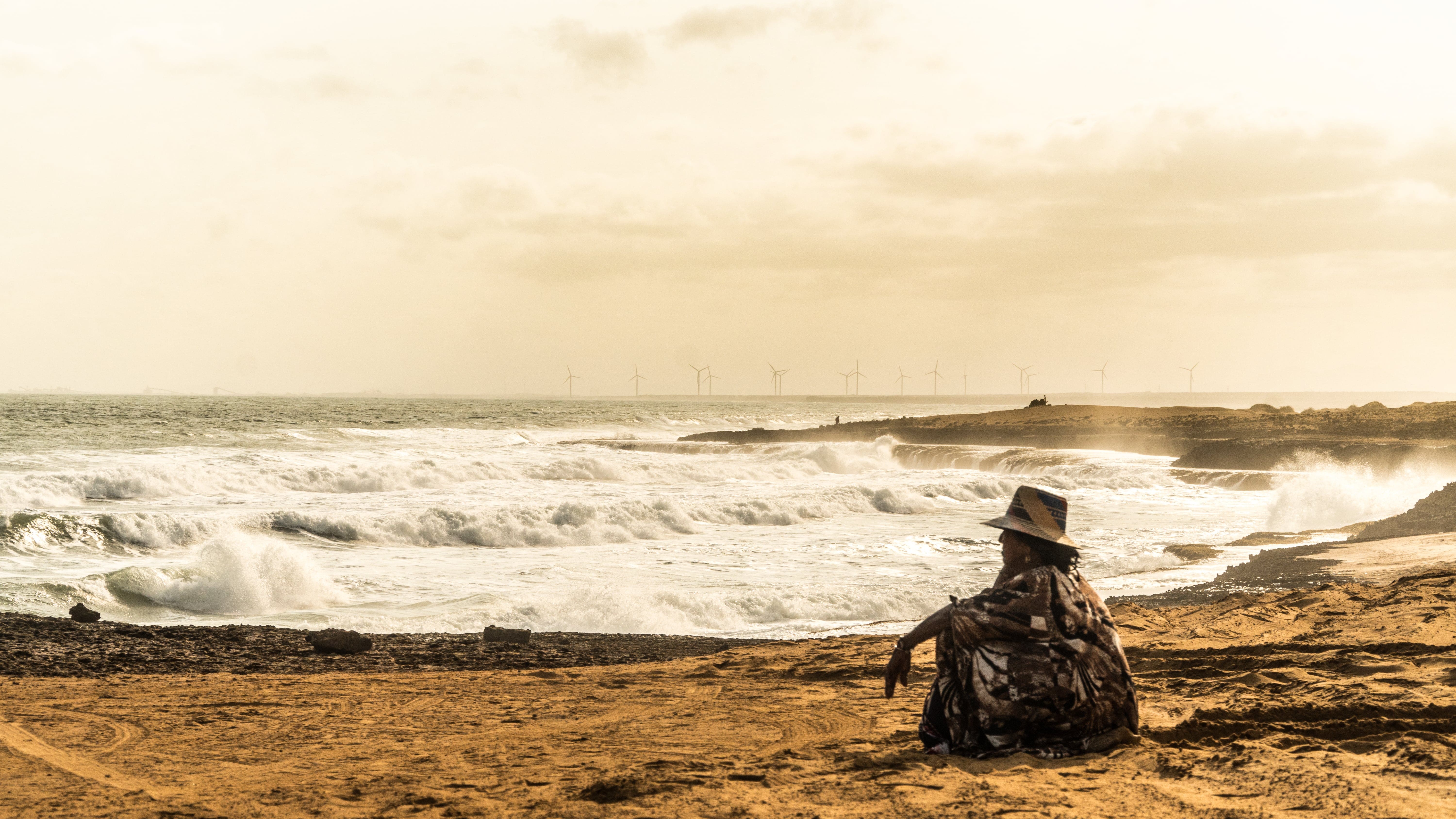 Georgina is looking away sitting on a beach next to a wind farm.