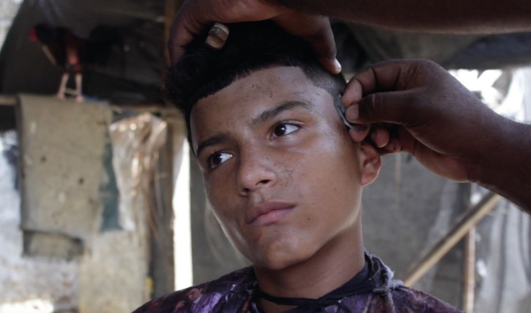 A teenager, Luis, looks away while a barber cuts his hair.