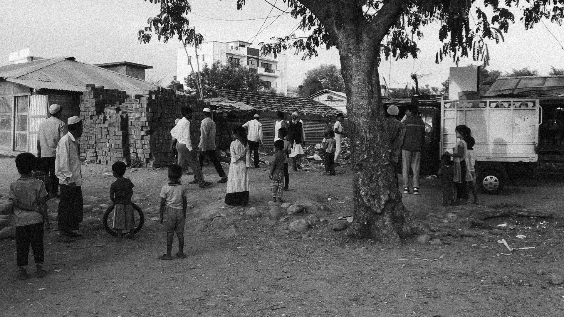 People standing by a tree with a refugee settlement in the background