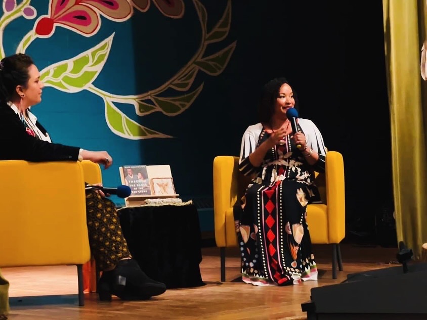 Maya Washington speaks into a microphone on stage across from AICHO (American Indian Community Housing Organization)Hannah Smith Arts and Culture Director