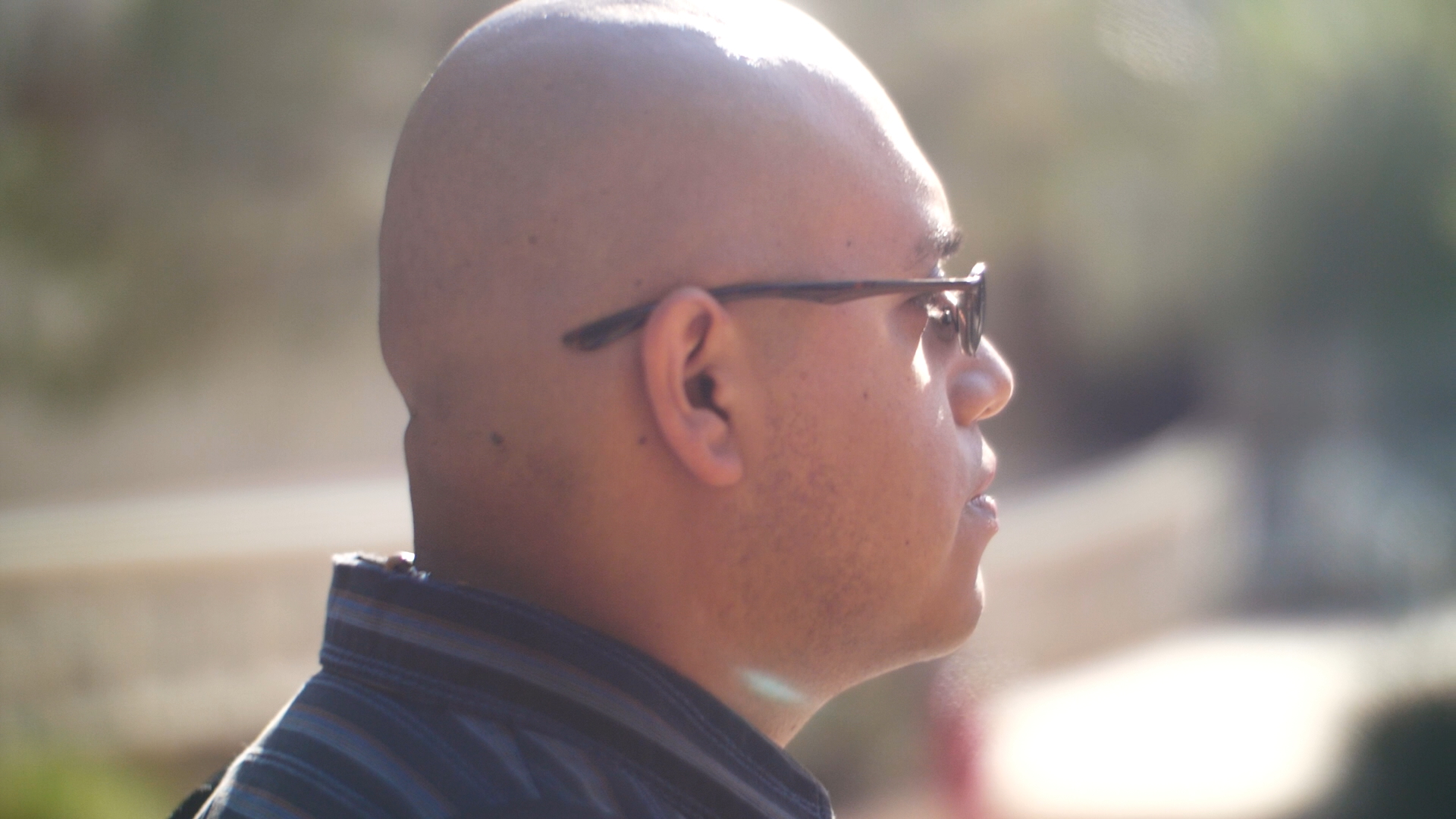 Side profile shot of a bald young man with glasses wearing a black collared shirt on a sunny day