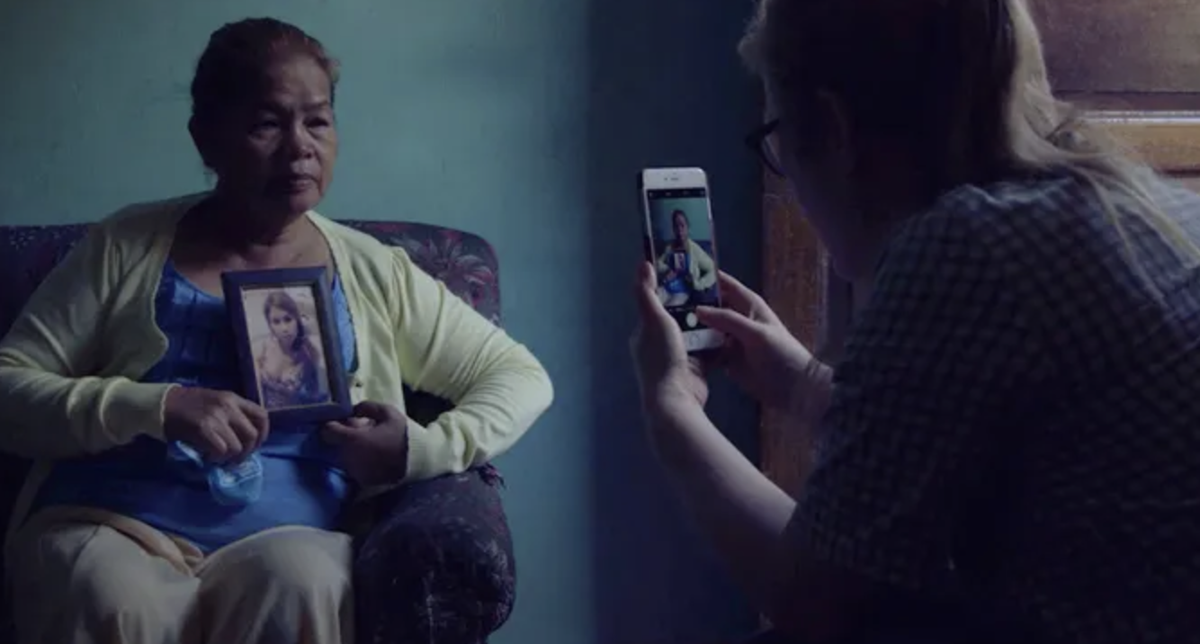 An older woman sits on a couch holding a picture frame as a woman sitting across from her takes a photo of her with her phone.