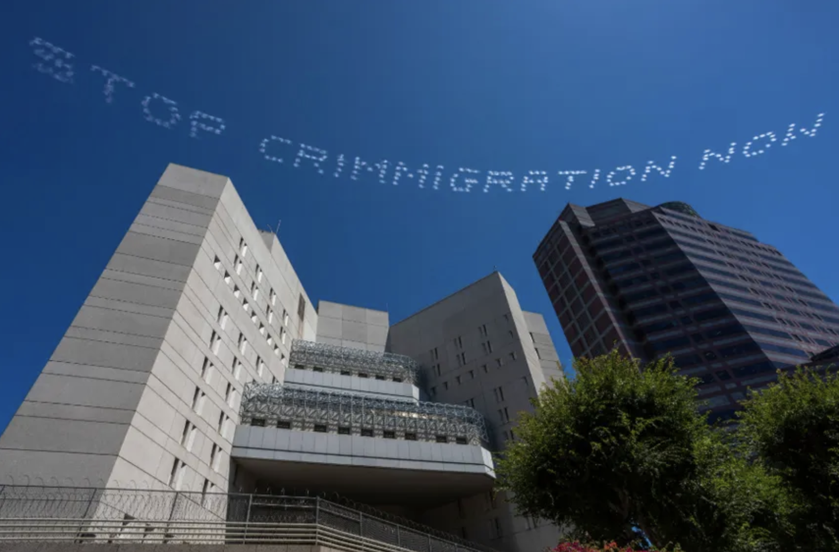 STOP CRIMMIGRATION NOW is typed as a cloud message against a blue sky above brutalist skyscrapers that serve as immigration prisons.