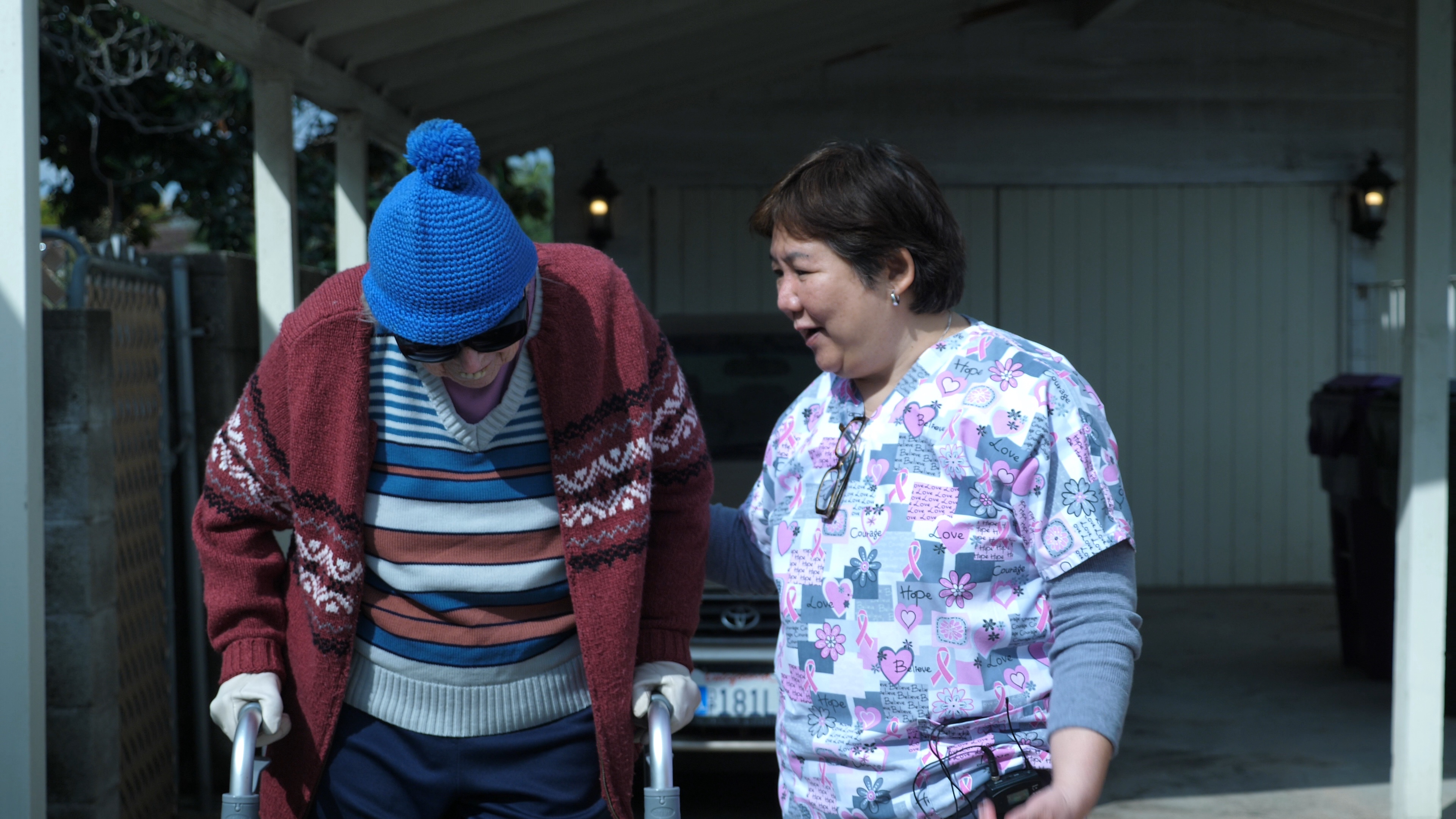 An elderly woman in a warm sweater and beanie looks straight down to the ground, smiling. Next to her is another woman wearing scrubs smiling back. They stand in front of a driveway.